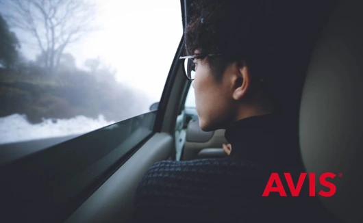 Person gazing out of a car window at a misty, snow-lined road, with Avis logo in the corner.