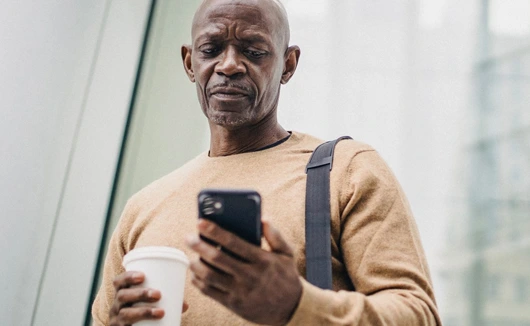 Man holding a coffee cup and checking his phone while walking indoors with a shoulder bag.