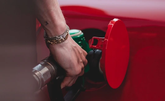 Close-up of a person’s hand inserting a fuel nozzle into a red vehicle at a petrol station.