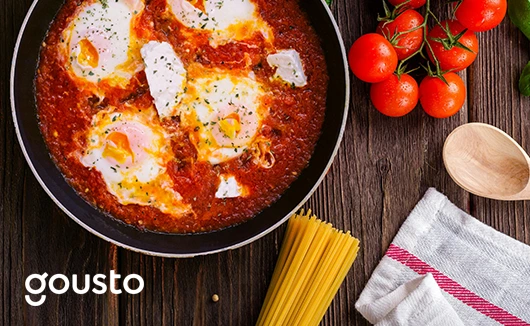 Shakshuka in a pan surrounded by fresh tomatoes, uncooked spaghetti, basil, and a tea towel, with Gousto branding.
