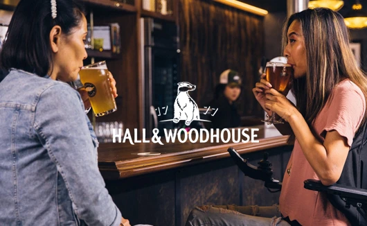 Two women enjoying beers at a Hall & Woodhouse pub bar, engaging in conversation.