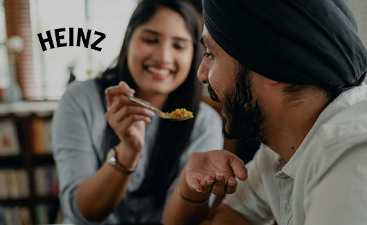 Smiling woman feeding a spoonful of food to a man, with Heinz logo in the corner.