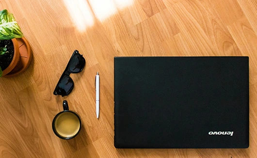 Flat-lay of a closed Lenovo laptop, sunglasses, pen, coffee cup, and potted plant on a wooden desk.