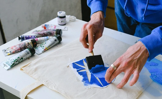 Person applying blue paint to a fabric surface using a stencil and squeegee, surrounded by paint tubes.