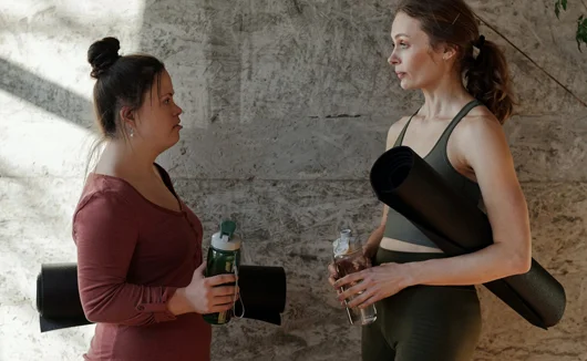 Two women holding water bottles and yoga mats, standing and talking in workout clothing.