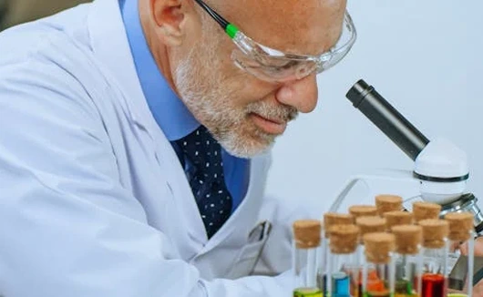 Male scientist in lab coat and safety goggles examining samples under a microscope, with colorful test tubes in the foreground.