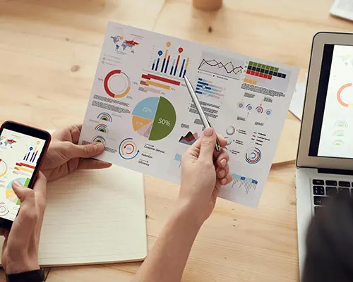 Hands holding a printed data report with charts, next to a phone and laptop displaying similar analytics.