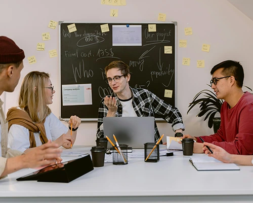 Team of young professionals brainstorming around a table with notes, coffee, and a chalkboard filled with ideas.