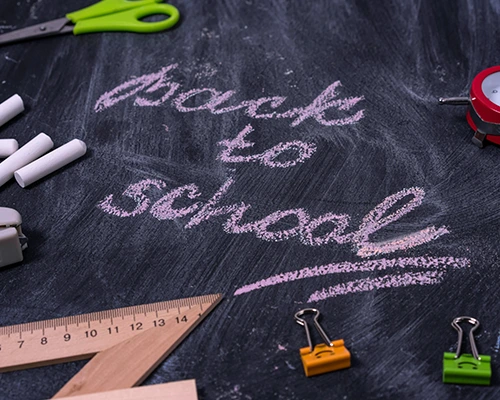 “Back to school” written in pink chalk on a blackboard, surrounded by stationery items like rulers, chalk, and paper clips.