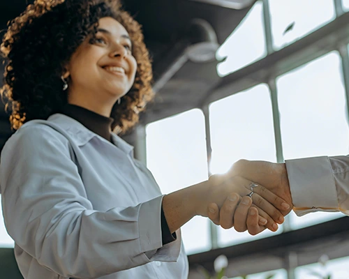 Smiling woman in professional attire shaking hands with another person in a sunlit office setting.