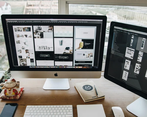 Dual-monitor setup displaying website design mockups on a wooden desk with stationery and a waving lucky cat ornament.