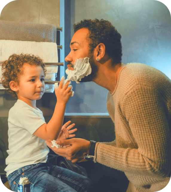 A young child applies shaving cream to an adult man's face in a bathroom