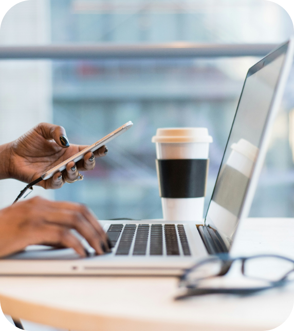 A person uses a laptop and holds a smartphone at a table. A coffee cup and eyeglasses are nearby, suggesting a focused, busy work environment.